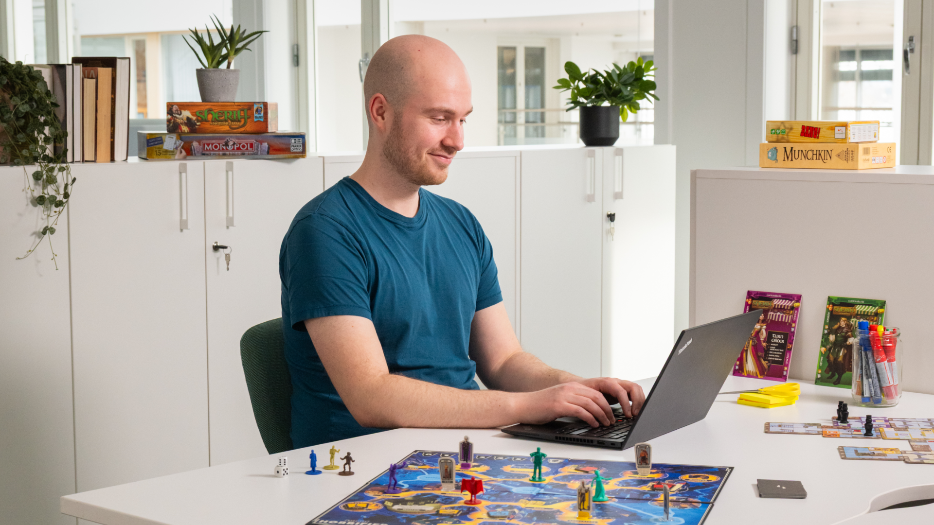 En mann sitter i et kontorlandskap foran en PC. Ved siden av ham står det et brettspill med spillfigurer